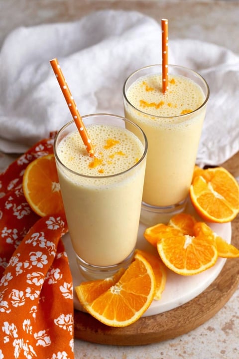 Two glasses of orange honey lassi with orange slices on a table.